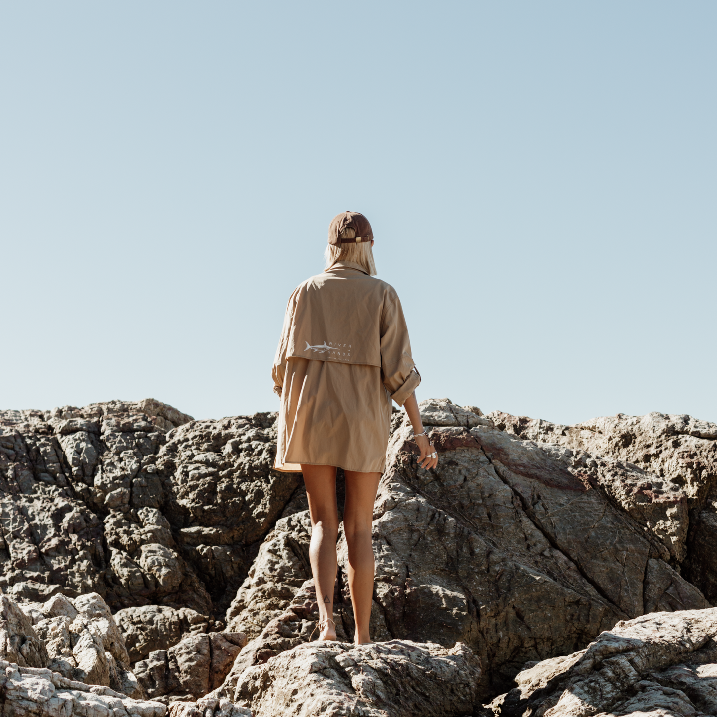 Back of a woman wearing Tan River Adventure Shirt showing the mesh back ventilation and she is climbing on rocks.