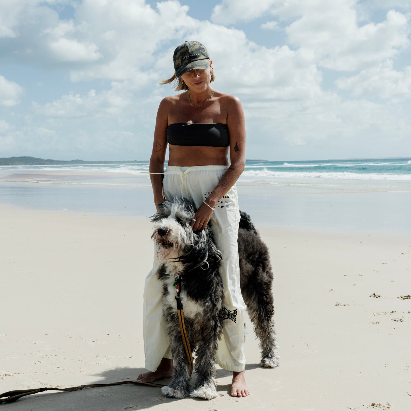 Woman standing on a beach with a large dog, wearing a black top and white pants and camo cap.