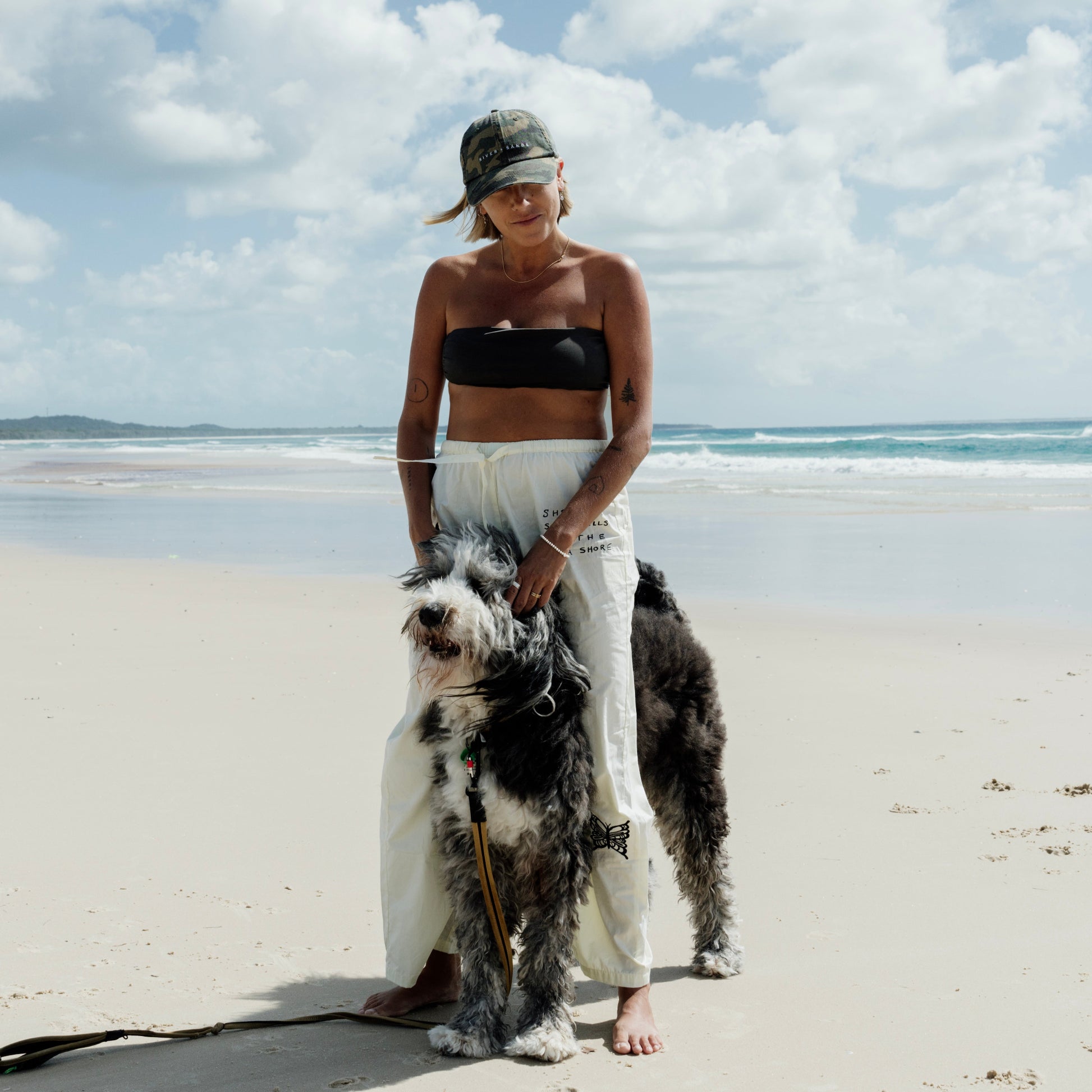 Woman standing on a beach with a large dog, wearing a black top and white pants and camo cap.