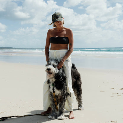 Woman standing on a beach with a large dog, wearing a black top and white pants and camo cap.