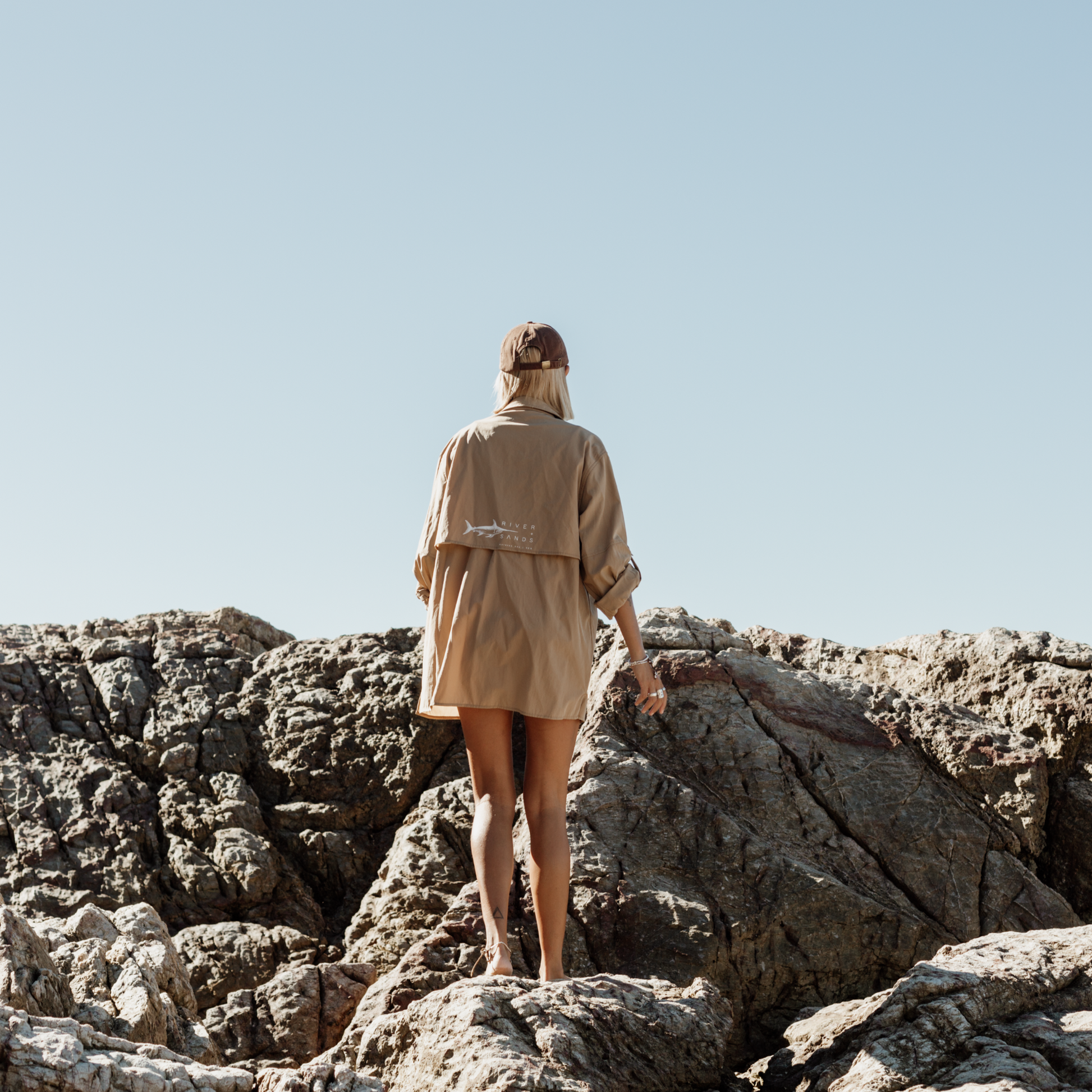 Back of a woman wearing Tan River Adventure Shirt showing the mesh back ventilation and she is climbing on rocks. 