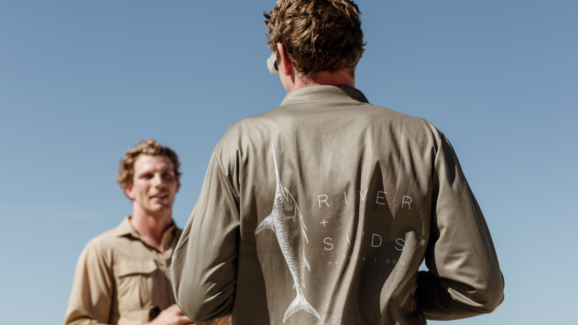 Two men wearing River and Sands shirts, one button up adventure shirt and the other a fishing polo, they are facing each other and smiling.