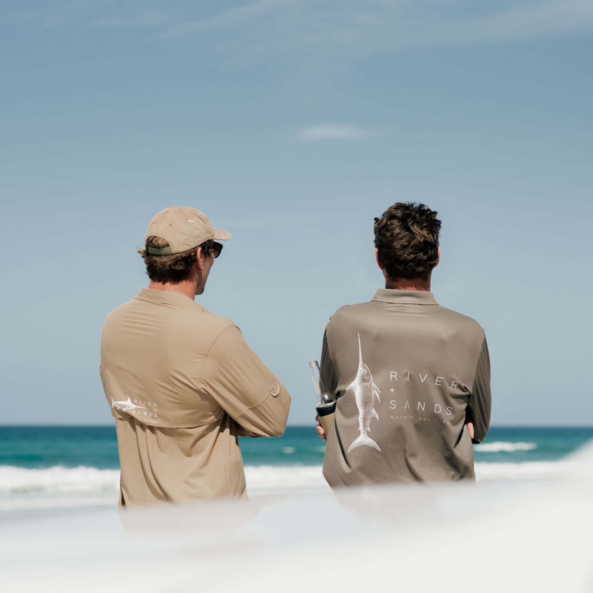 Two men wearing a tan river adventure shirt and a sands fishing shirt, they are on the beach looking out to the water.