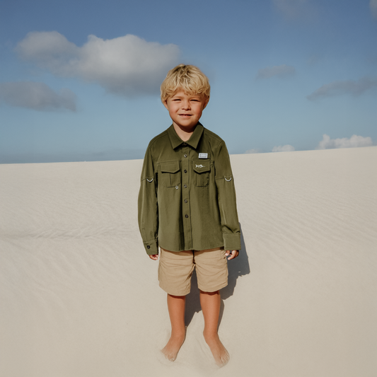 Young boy standing on a sandy dune wearing an olive green River and Sands shirt and beige shorts.